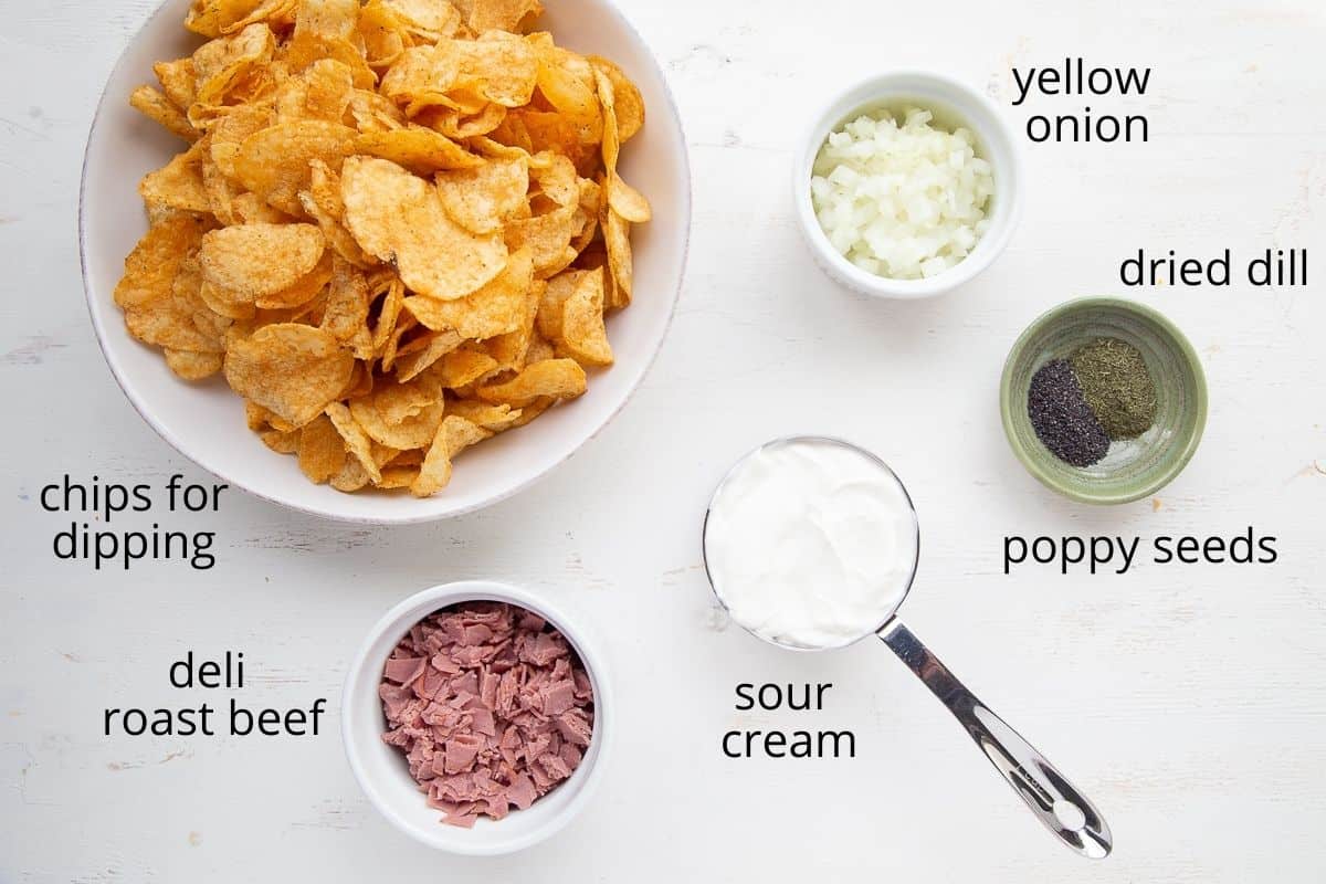 potato chips, chopped deli beef, sour cream, yellow onion, dried dill, and poppy seeds on a white table.