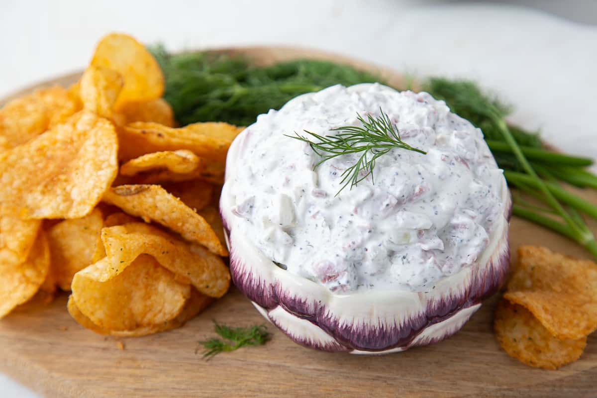 beef chip dip in a small round bowl on a wooden board, surrounded by potato chips and fresh dill.