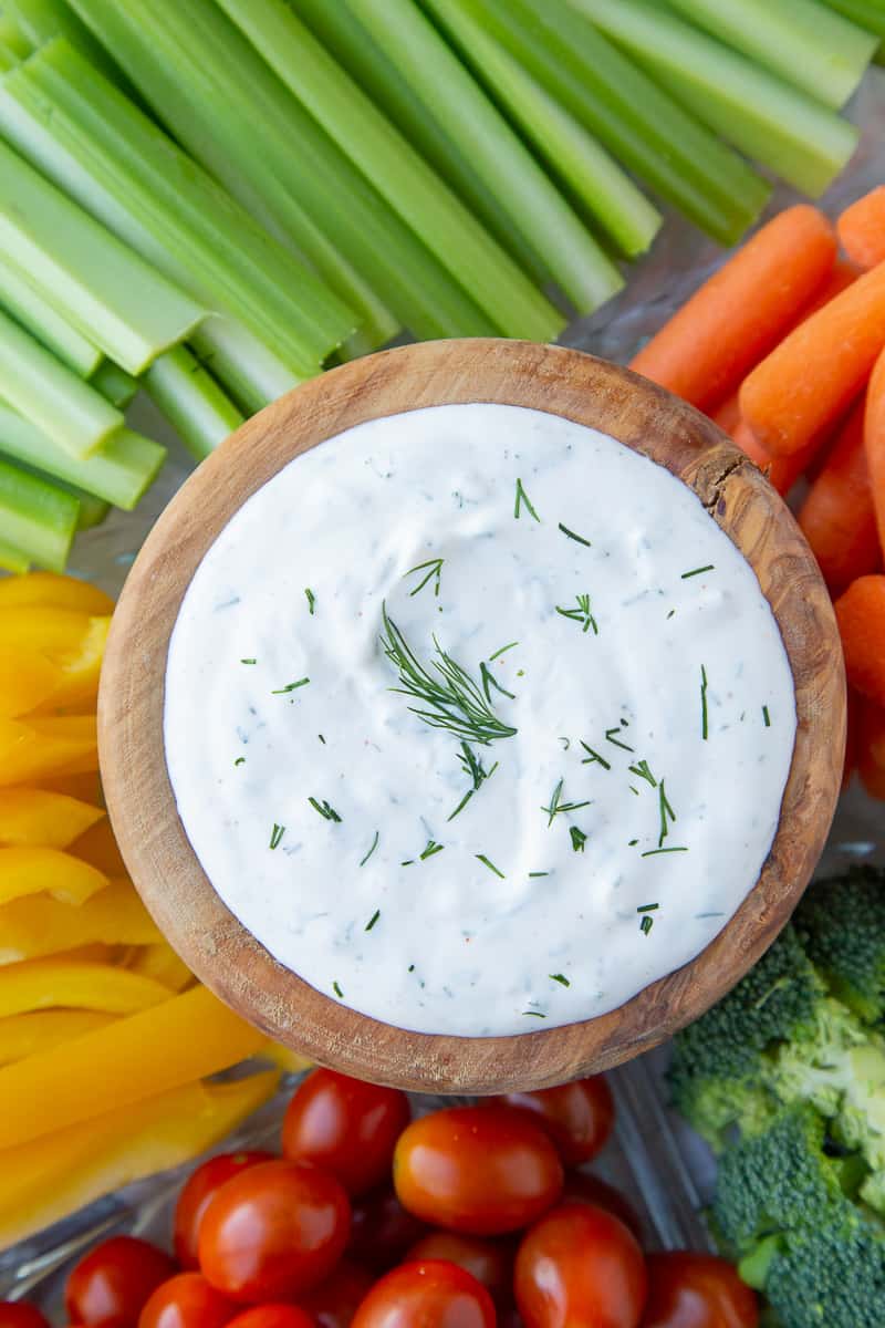 wooden bowl full of sour cream dill dip surrounded by celery, carrots, broccoli, tomatoes, and bell pepper slices.