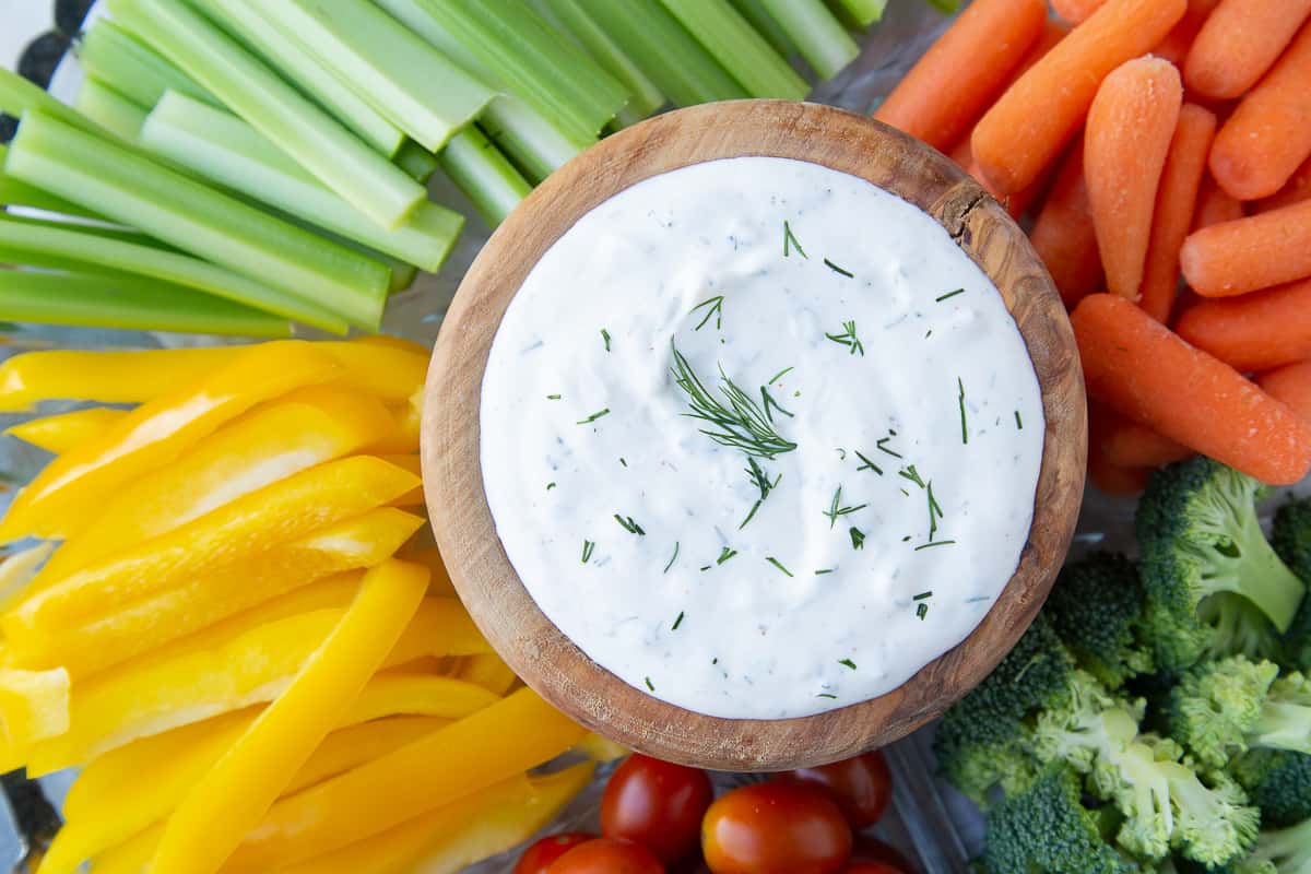 white dip in a wooden bowl, garnished with fresh dill and surrounded by fresh raw veggies.