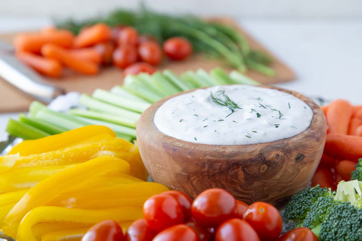 sour cream dill dip in a wooden bowl surrounded by fresh veggies, with a cutting board and knife with more veggies in the background.