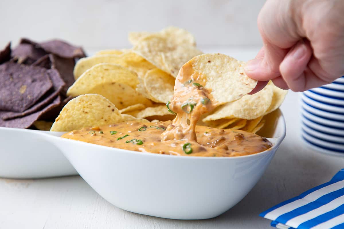a hand scooping up nacho cheese dip with a yellow corn tortilla chip.