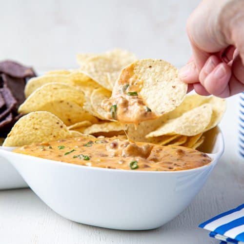 hand dipping a chip into nacho cheese dip in a white bowl.