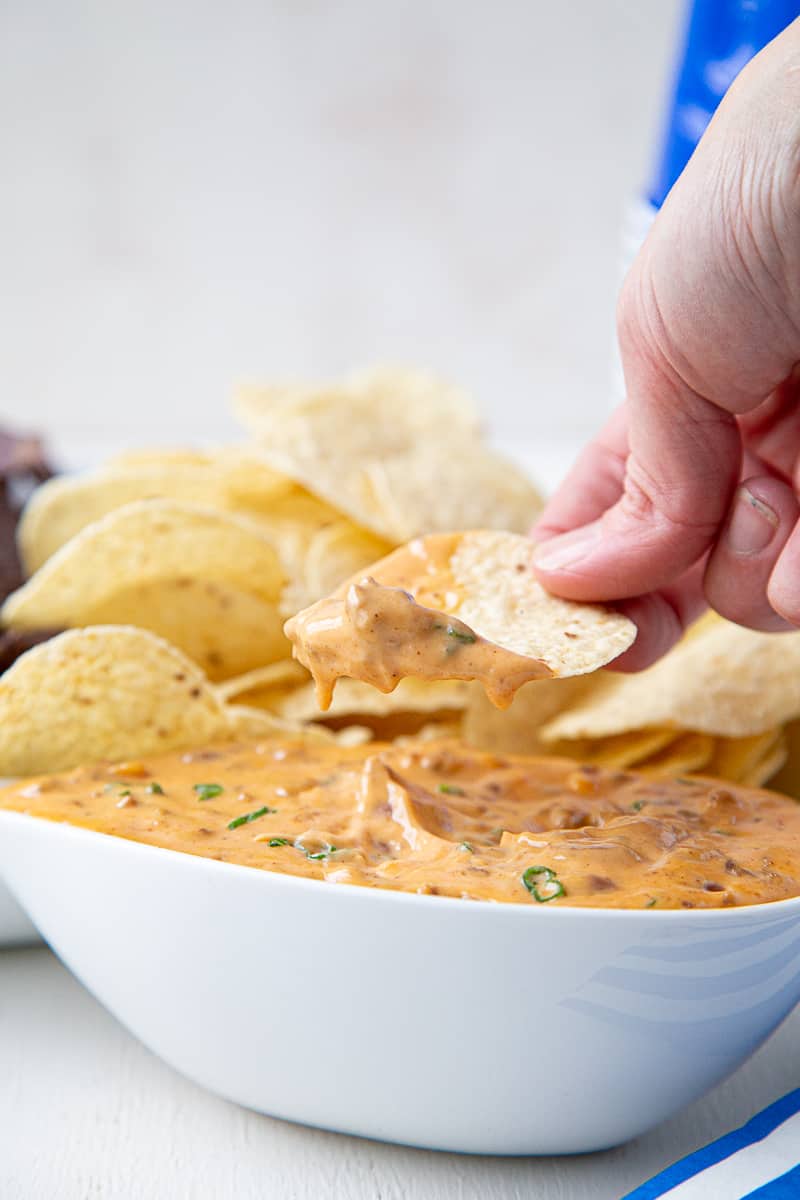 hand dipping a tortilla chip into loaded nacho cheese dip in a white bowl.