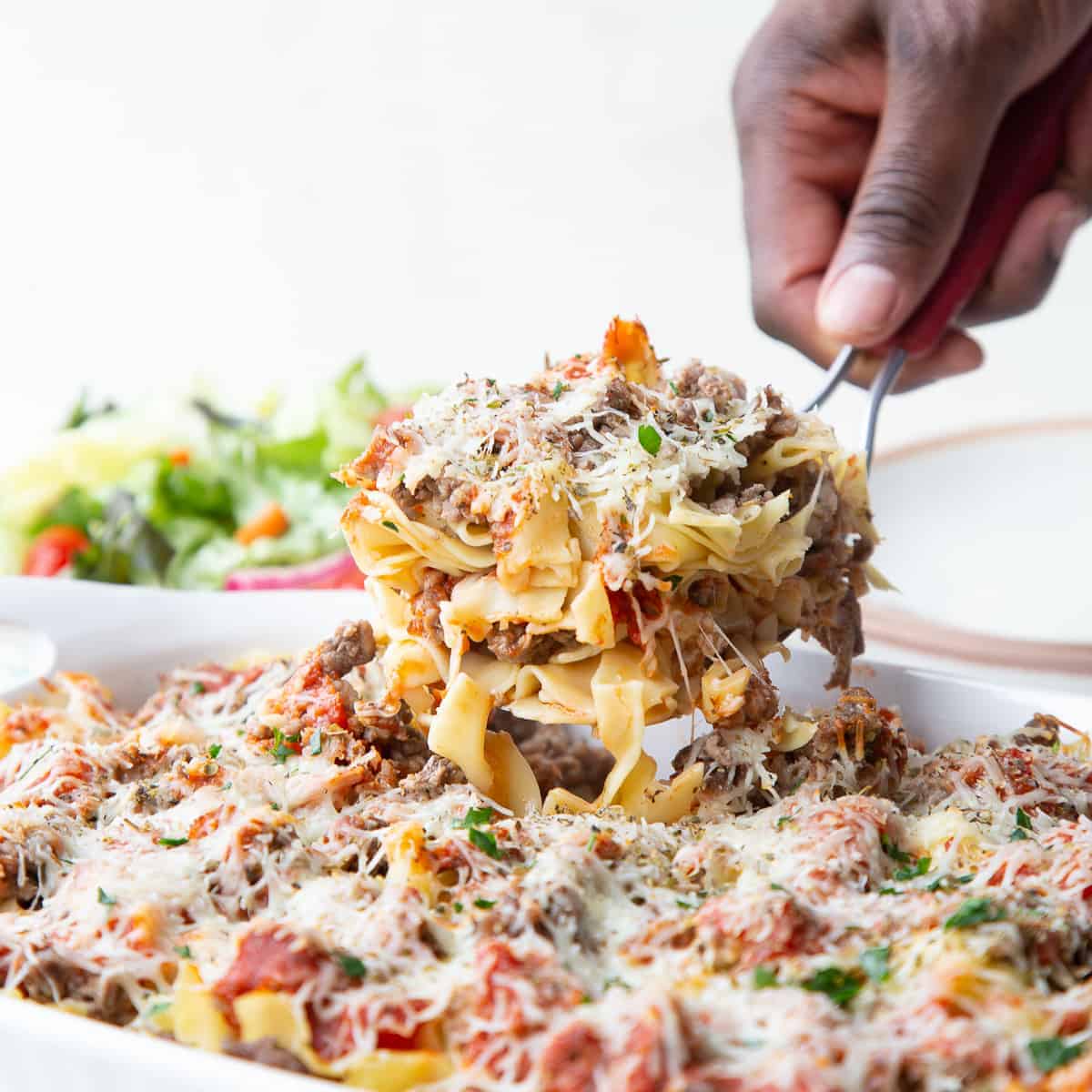 hand lifting a scoop of pizza casserole from a white casserole dish.