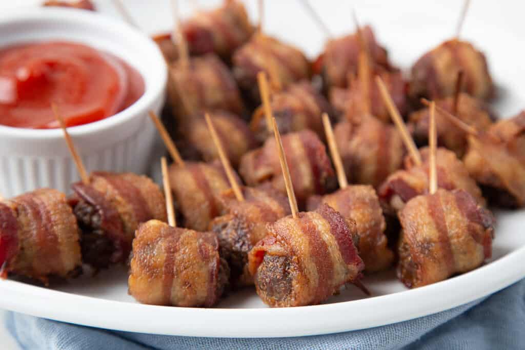 white oval platter filled with bacon wrapped meatballs, with a small bowl of ketchup on the side.