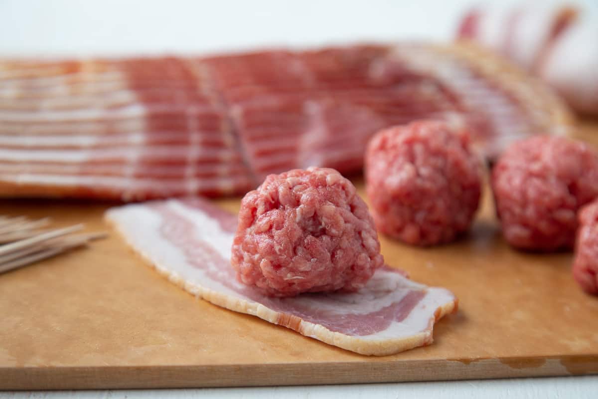 raw meatball sitting on top of half a slice of uncooked bacon, with more bacon and raw meatballs in the background.