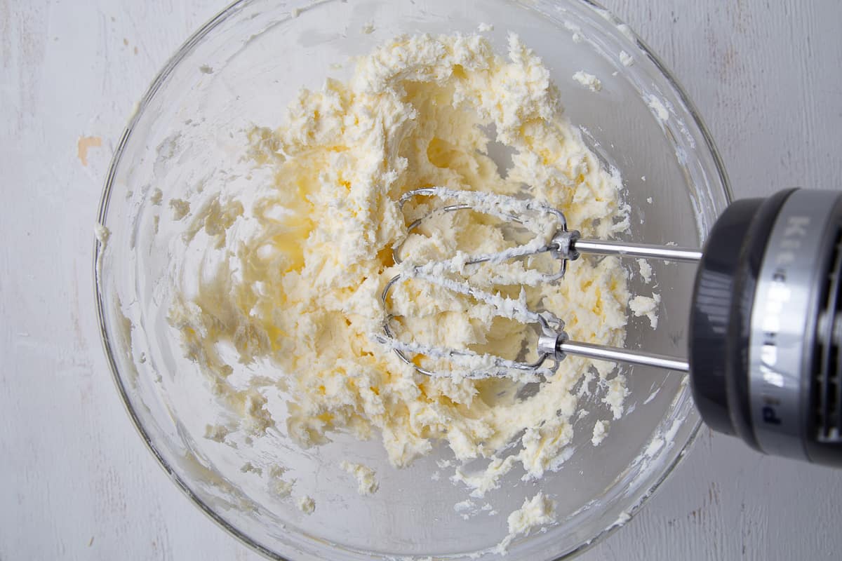 handheld mixer mixing butter and cream cheese in a glass mixing bowl.