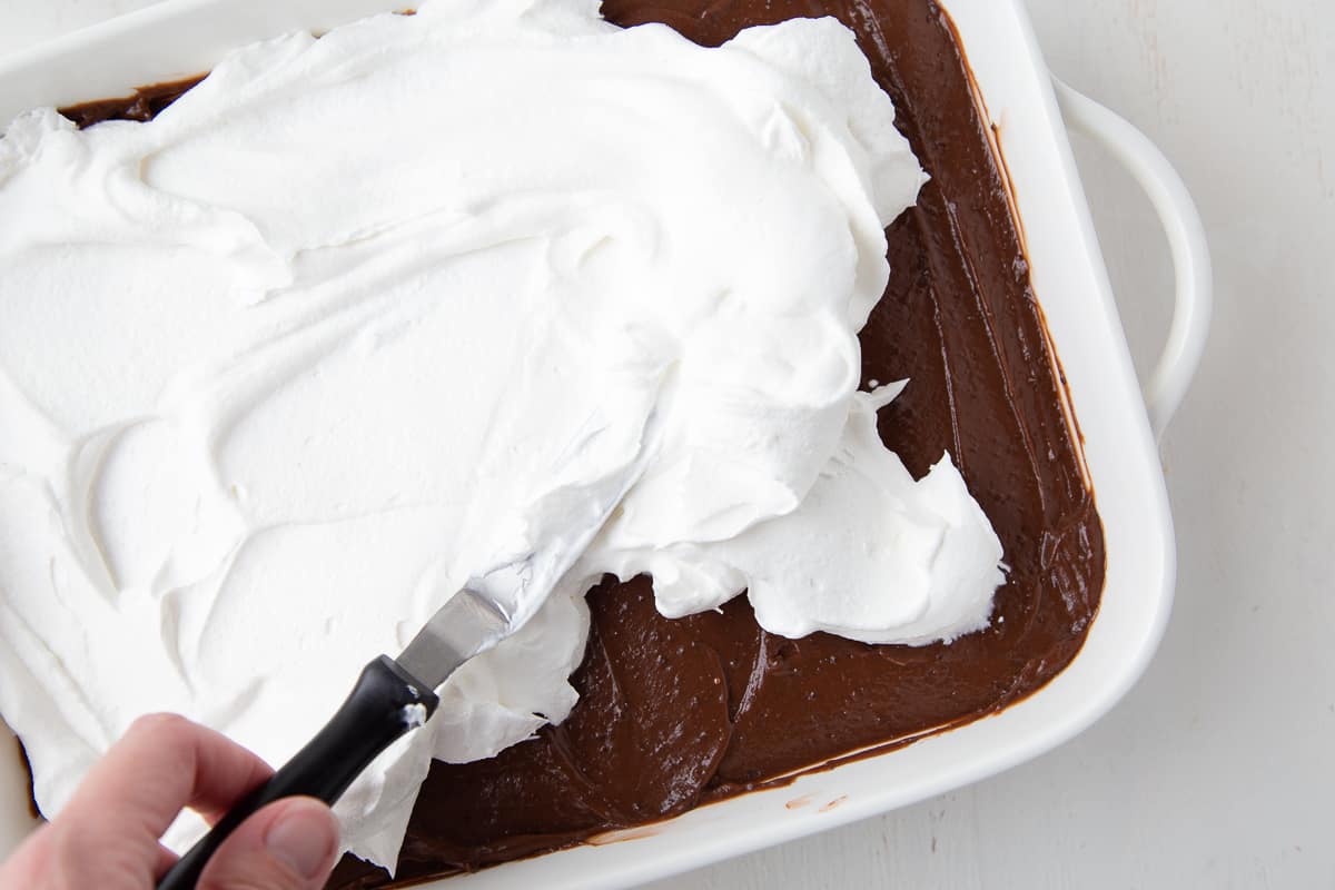 hand using an offset spatula to spread whipped cream on top of chocolate pudding in a casserole dish.