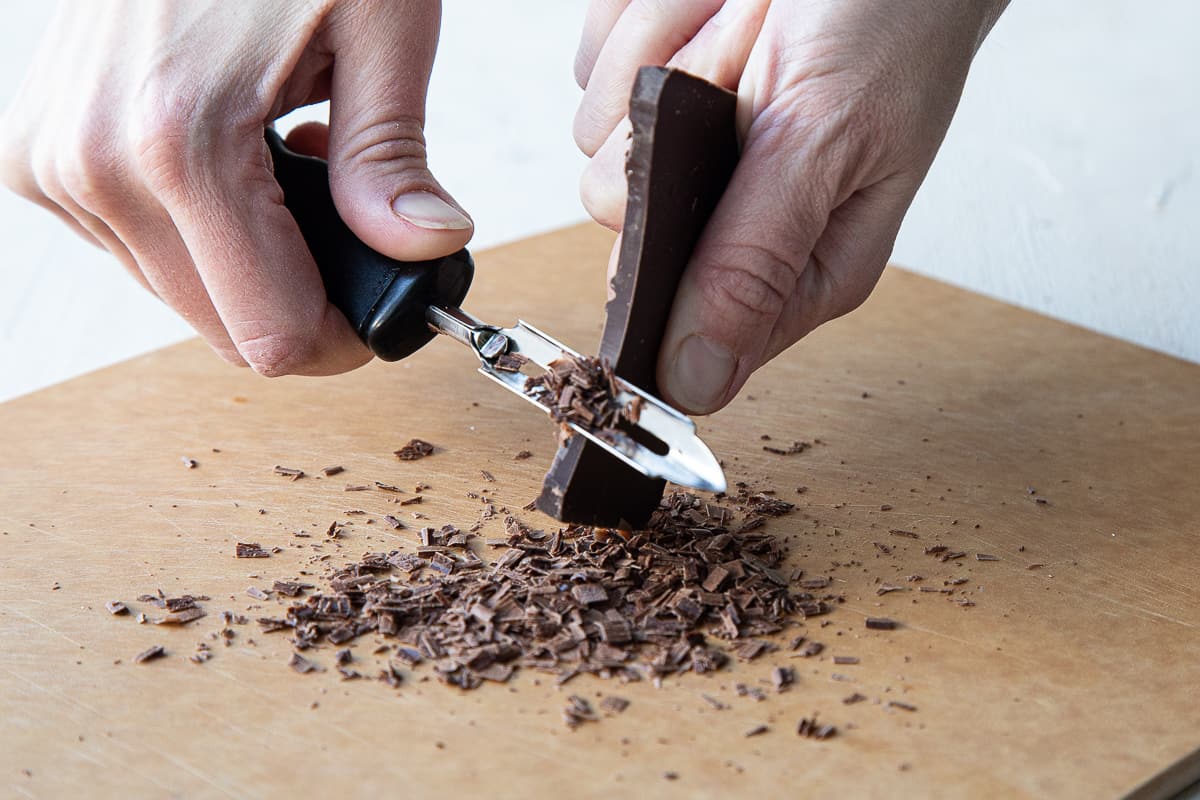 hands using a vegetable peeler to create chocolate shavings from a chocolate bar.