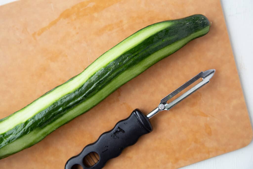 partially peeled cucumber on a wooden board next to a vegetable peeler.