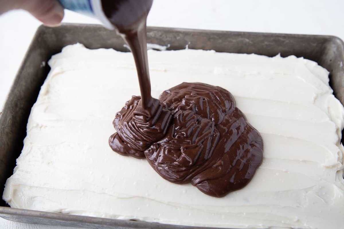 hand pouring a can of chocolate frosting over a cake in a metal pan.