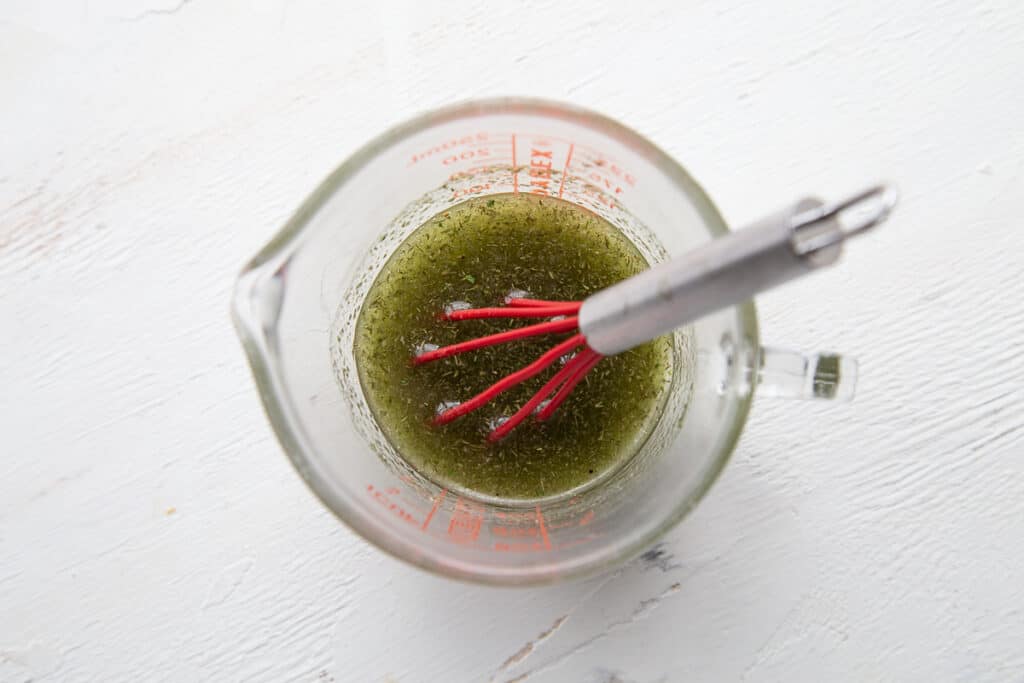 measuring cup full of oil and herbs, with a mini whisk in it.