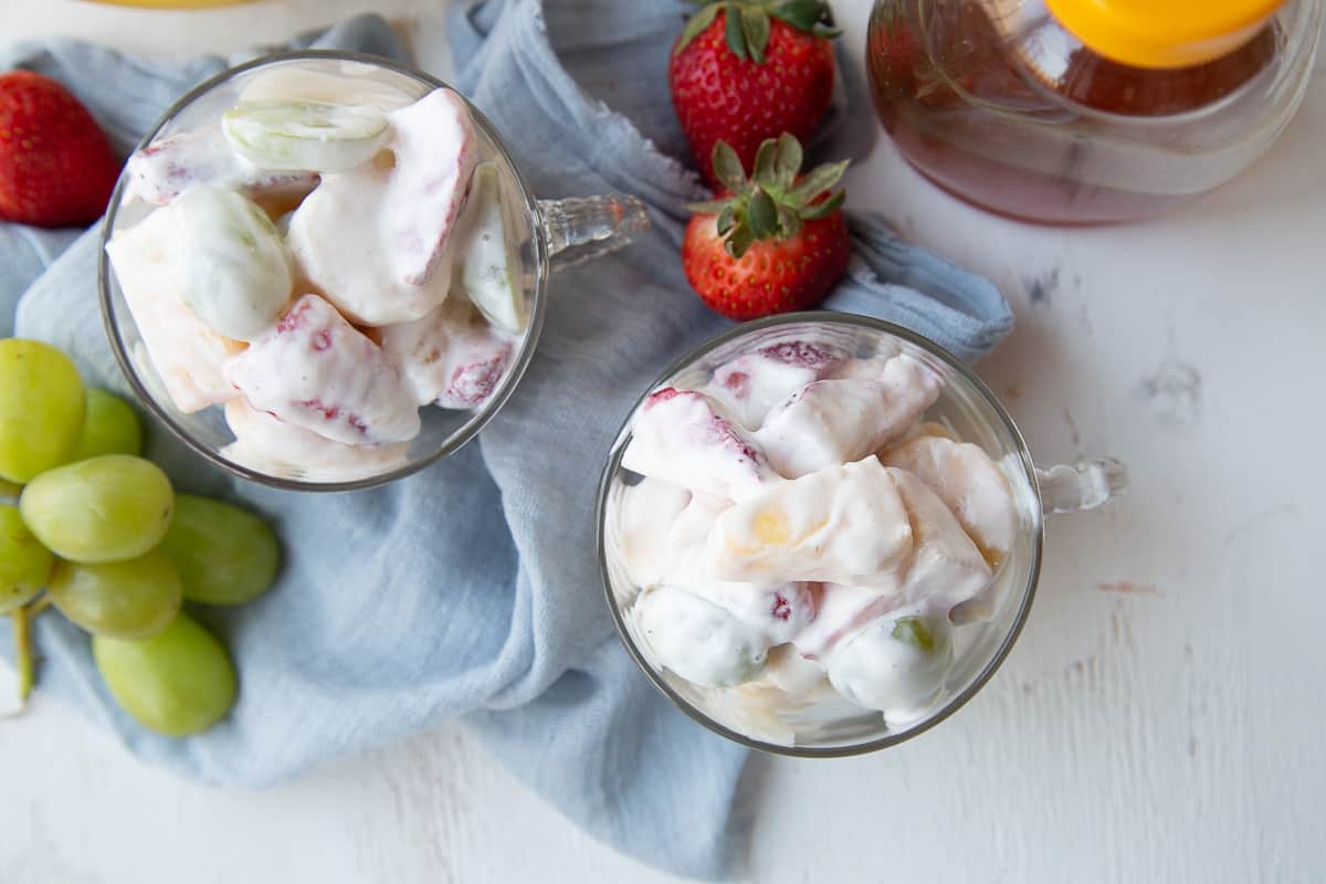 two glass cups full of creamy fruit salad, surrounded by fresh fruit, sitting on a blue dish towel.