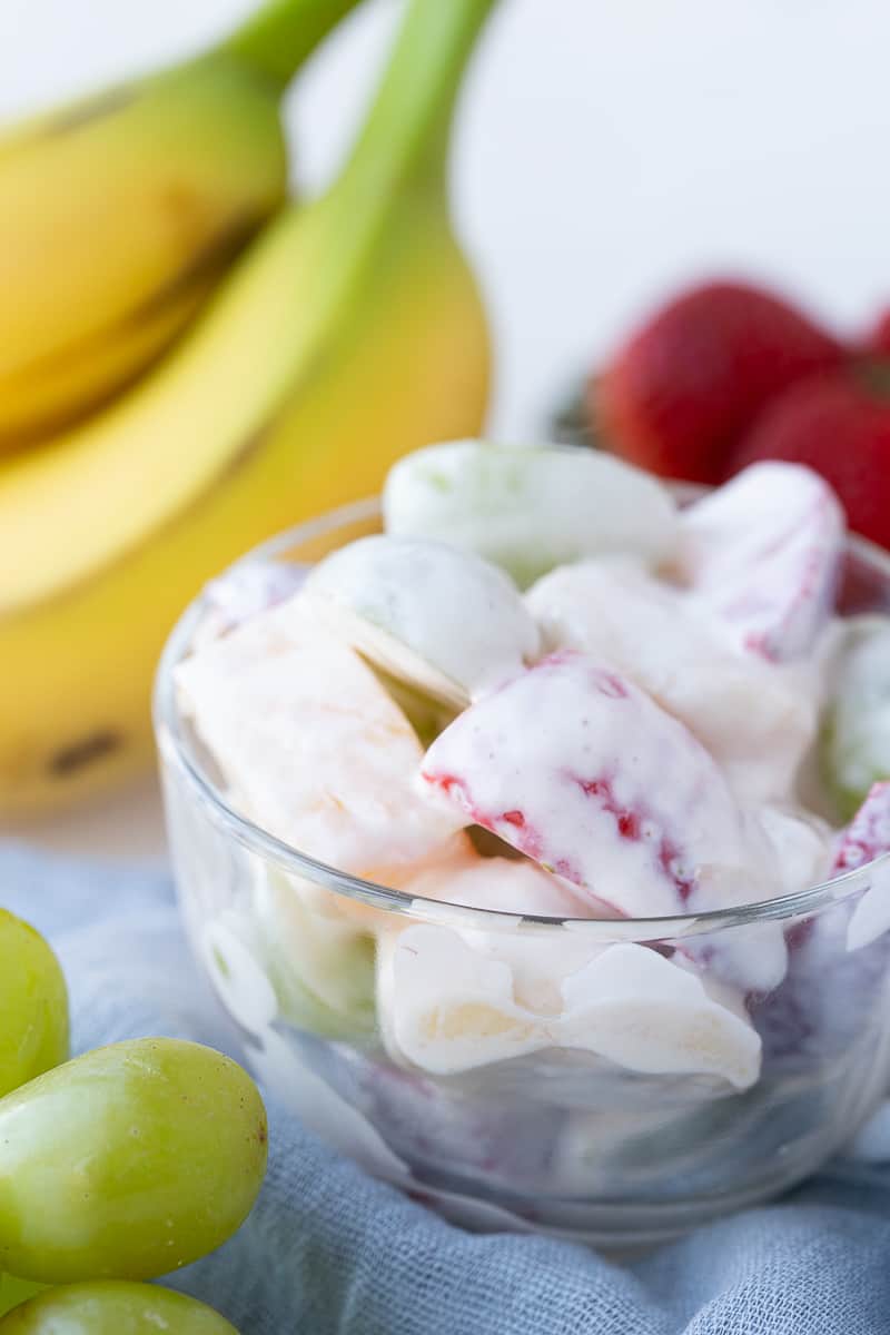 creamy fruit salad in a glass cup next to whole bananas and strawberries and grapes.