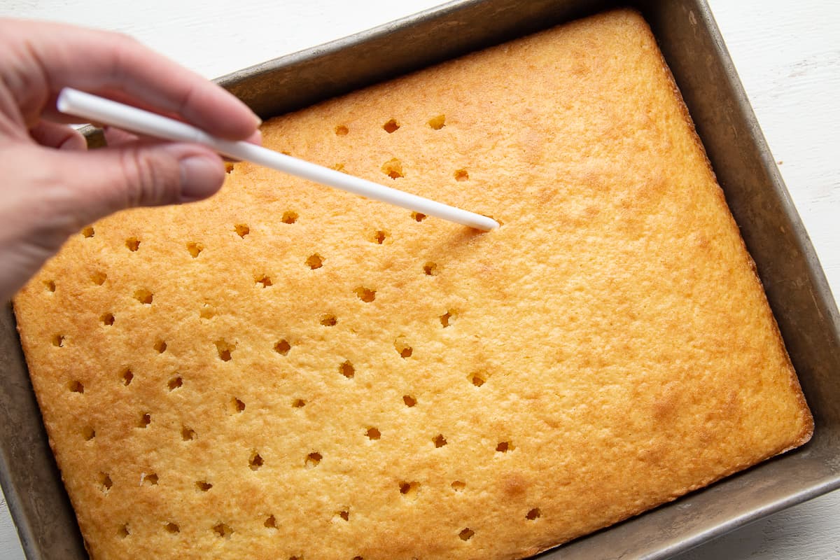 hand using a straw to poke holes in the top of a white cake.