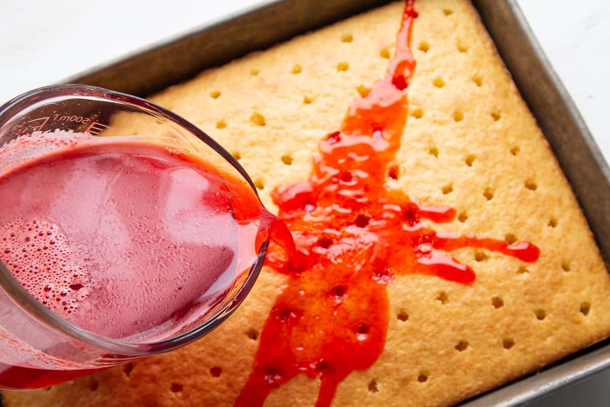 glass measuring cup pouring red liquid over a white cake. 