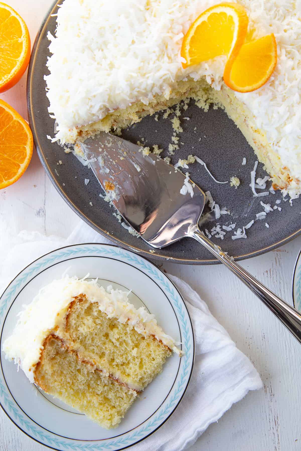 slice of orange layer cake on a blue and white plate next to a whole orange cake with orange peel garnish on a gray plate.