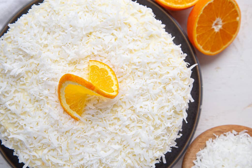 coconut topped cake, topped with a twisted orange slice with a bowl of coconut and a halved orange for garnish.