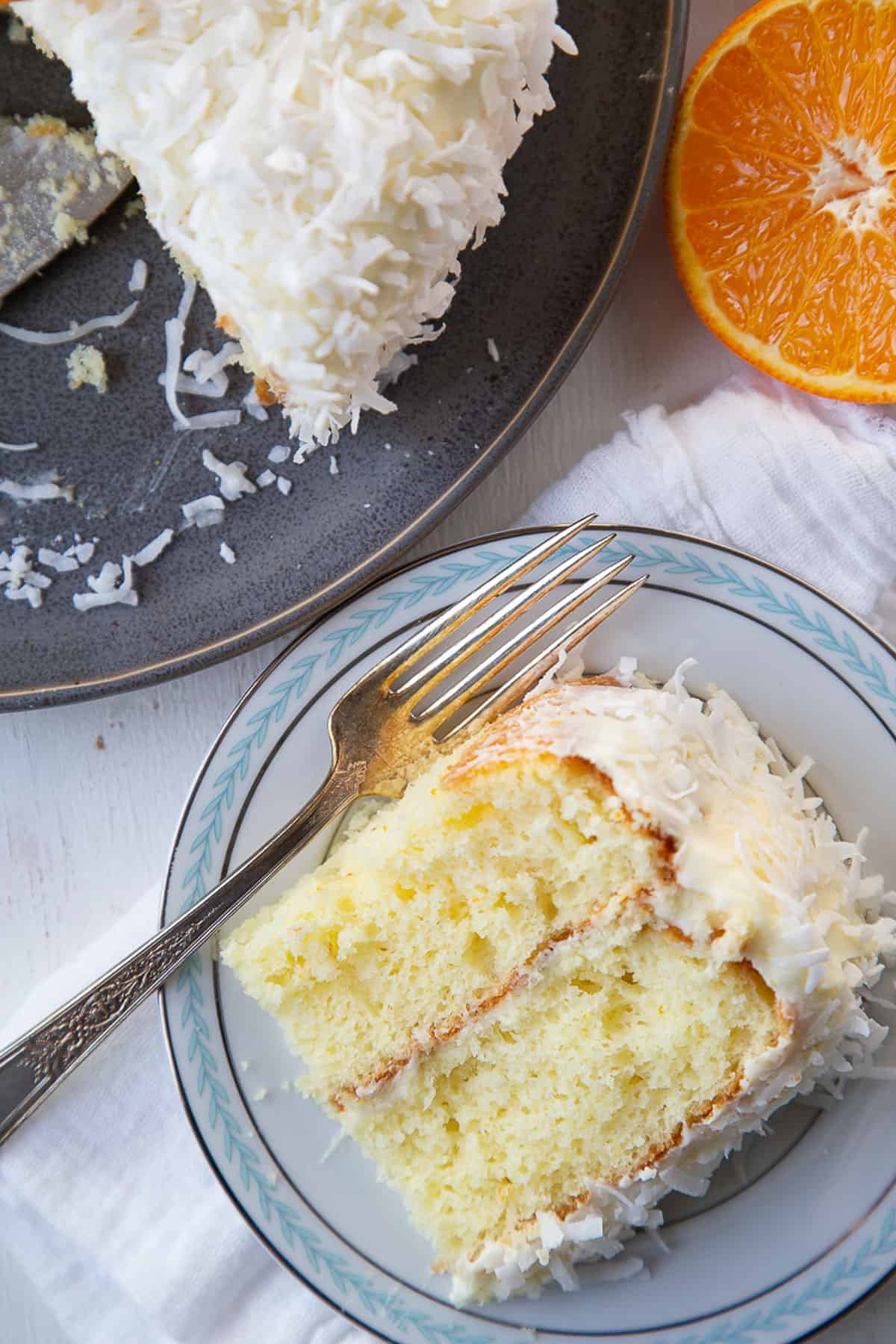 slice of orange layer cake with coconut frosting on a plate next to a whole cake.