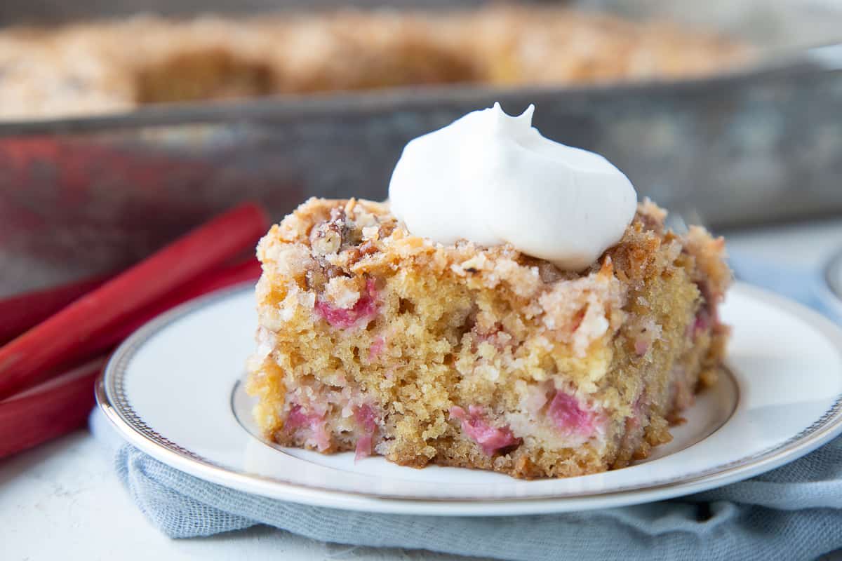 slice of rhubarb cake on a white plate, topped with whipped cream with a metal pan of cake in the background.
