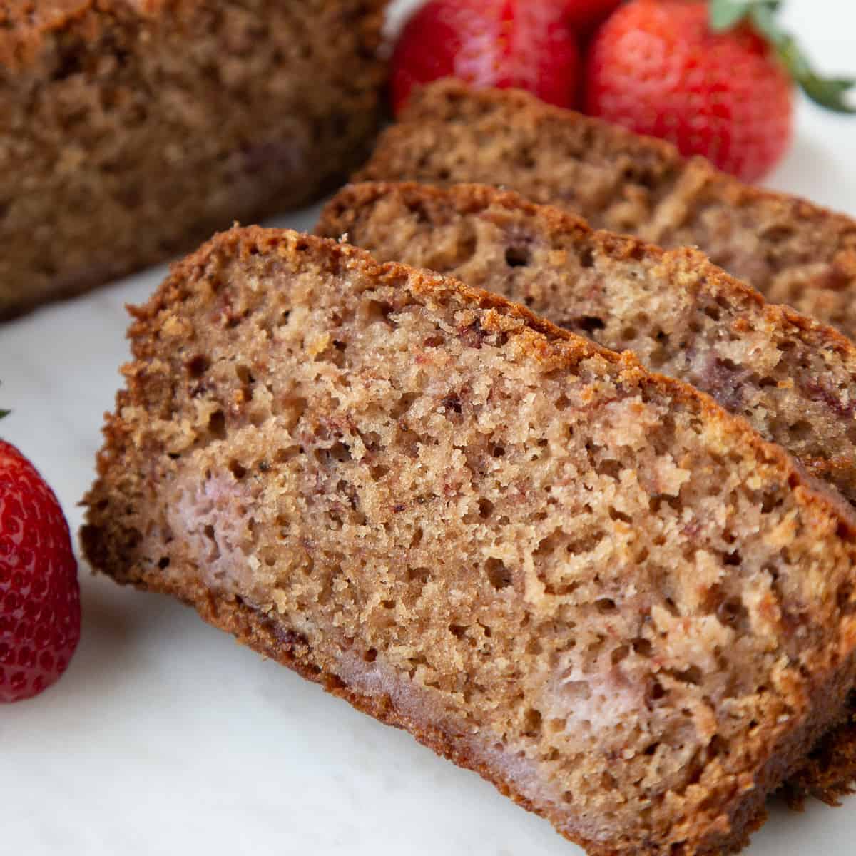 slices of strawberry bread on a white marble platter with fresh strawberries in the background.