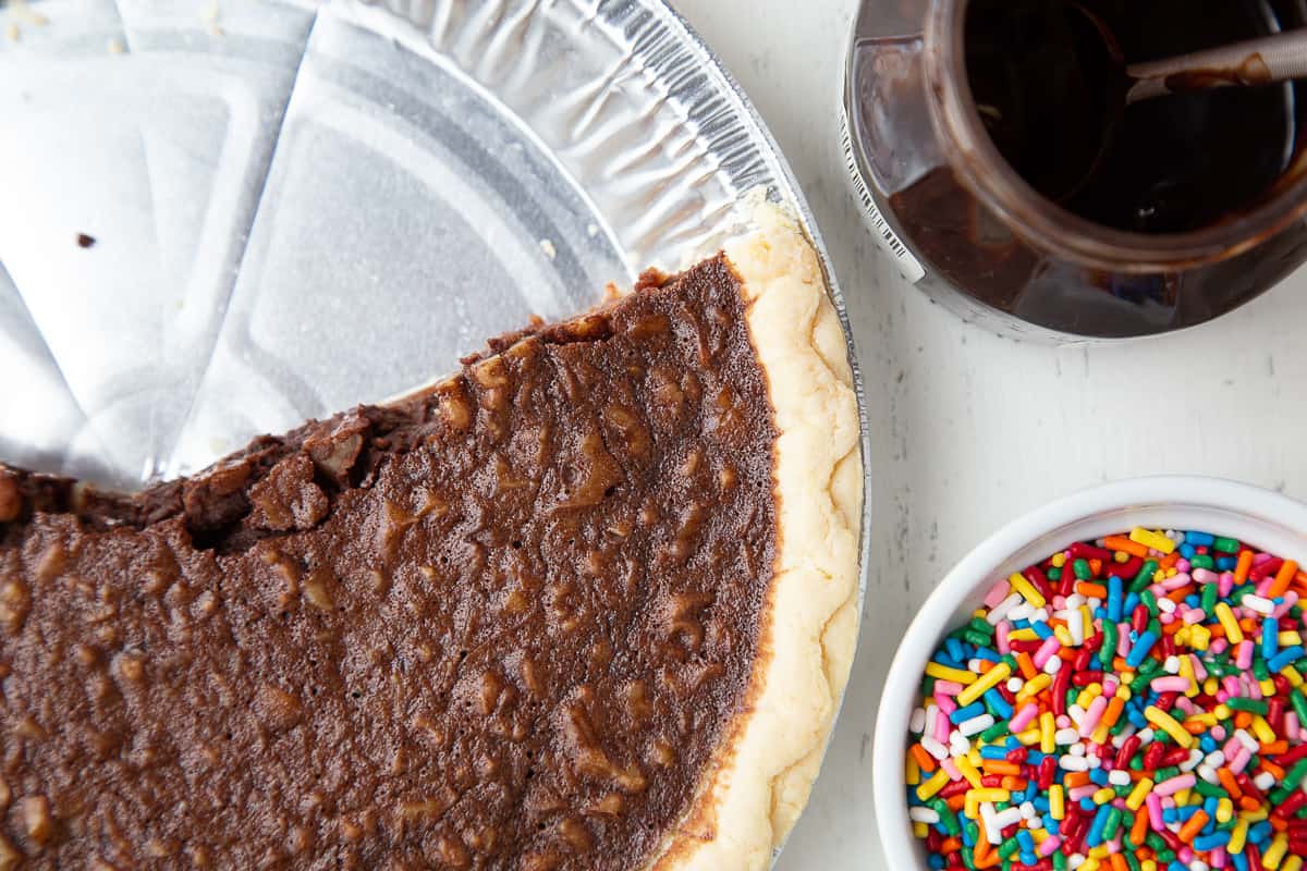 brownie pie with slices taken out, next to chocolate sauce and rainbow sprinkles.