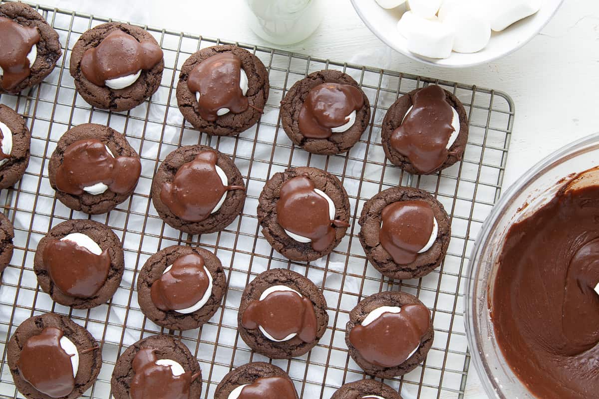 chocolate marshmallow cookies on a wire rack next to a bowl of fudge icing.