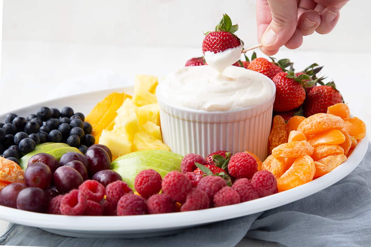 hand dipping a strawberry into fruit dip, sitting on a platter of fresh fruit.