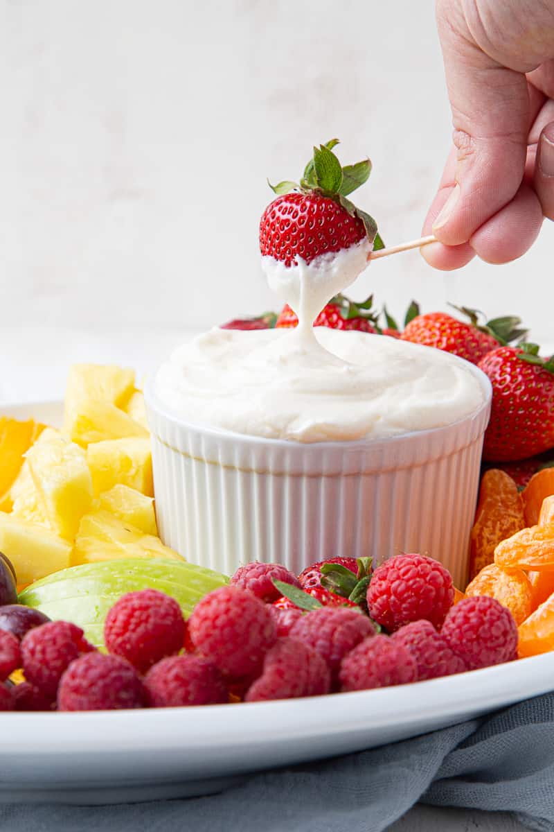 hand dipping a strawberry into fruit dip in a white bowl, surrounded by cut fruit.