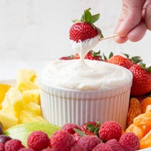 hand dipping a strawberry into a white bowl of fruit dip, surrounded by fruit.
