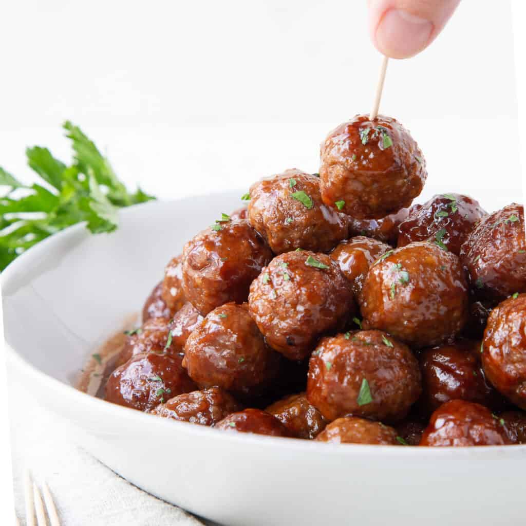 hand picking up a meatballs with a toothpick from a white bowl full of crockpot bbq meatballs.