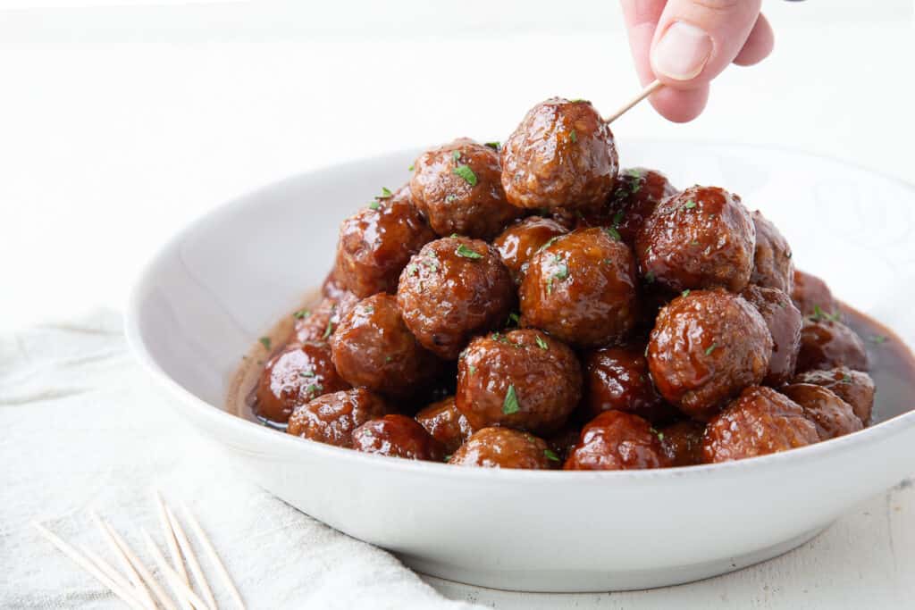 hand picking up a meatballs from a white platter of bbq meatballs.