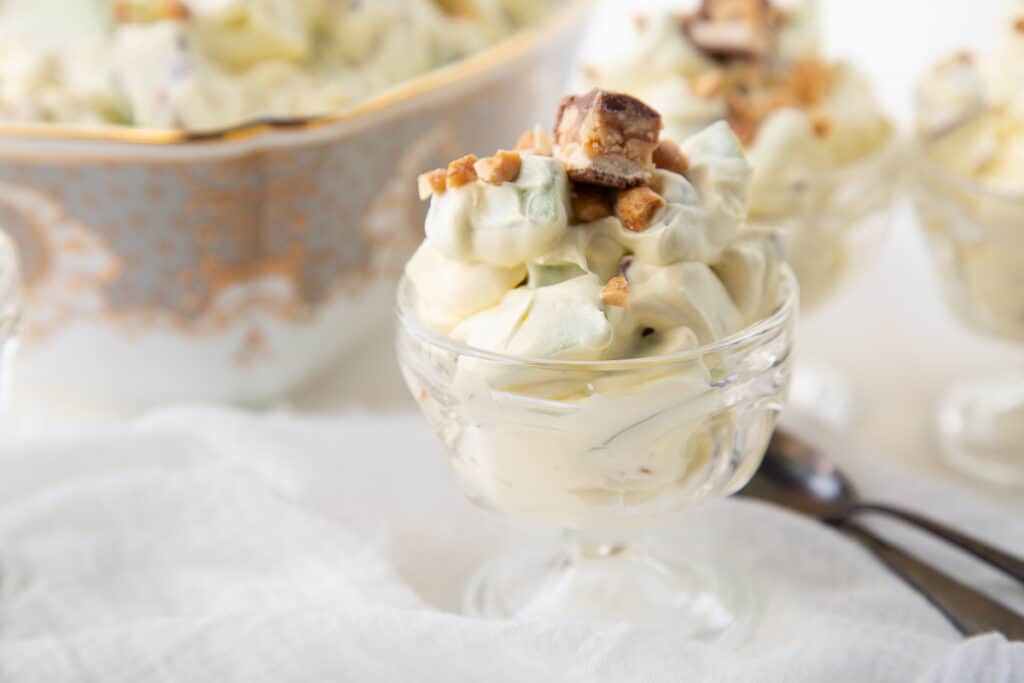 parfait glass of snickers salad next to two spoons and a big bowl of snickers salad.