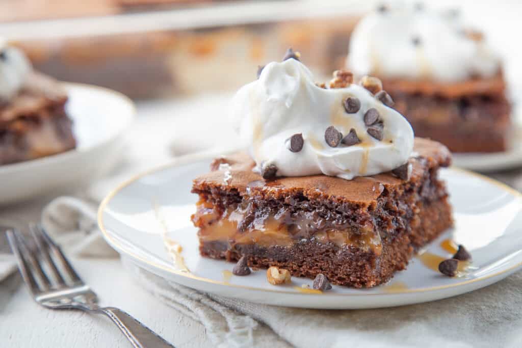 turtle cake slice on a white plate, topped with whipped cream.