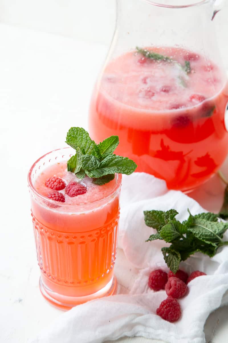 glass of raspberry lemonade garnished with mint next to a pitcher of more lemonade.