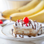 slice of banana split cake drizzled with chocolate sauce and topped with a maraschino cherry.