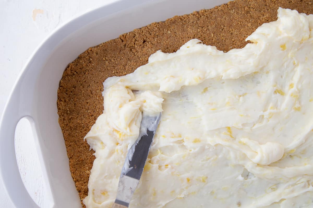 offset spatula spreading cream cheese mixture over a graham cracker crust in a white pan.