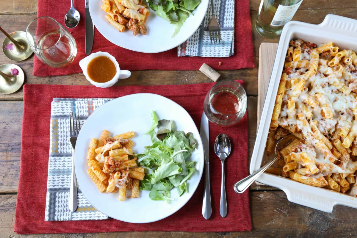 table set with red placemats, white plates, a casserole dish of baked ziti, and plates filled with ziti and salad.