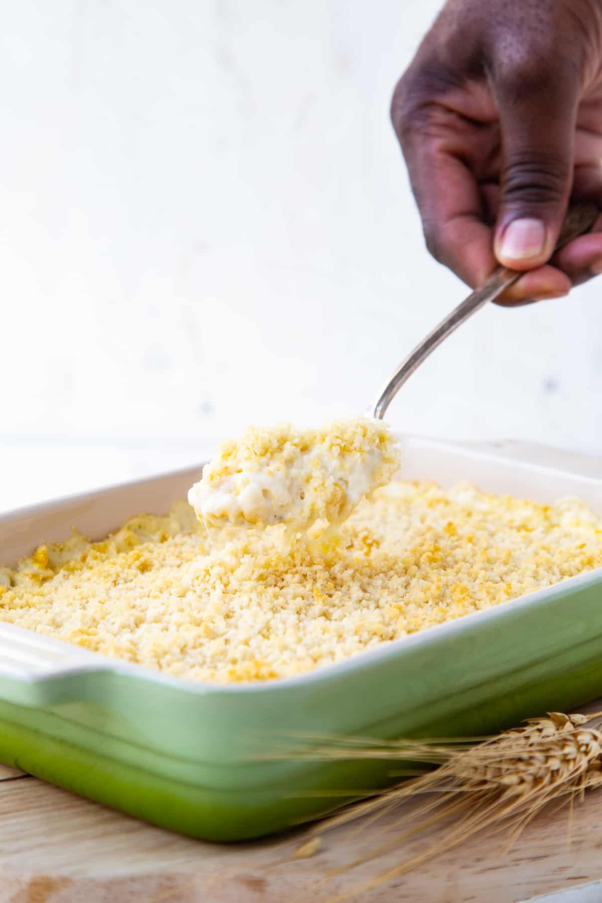 hand lifting cheesy corn casserole out of a green casserole dish.