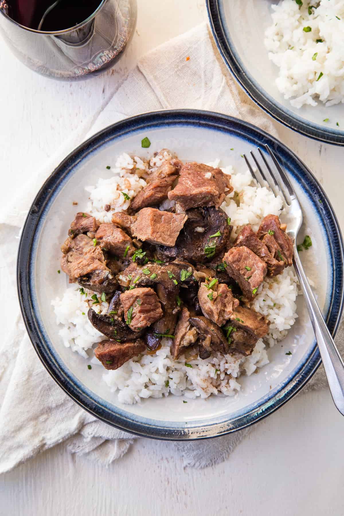 beef stew with rice on a blue rimmed plate on a dinner table with a glass of red wine.