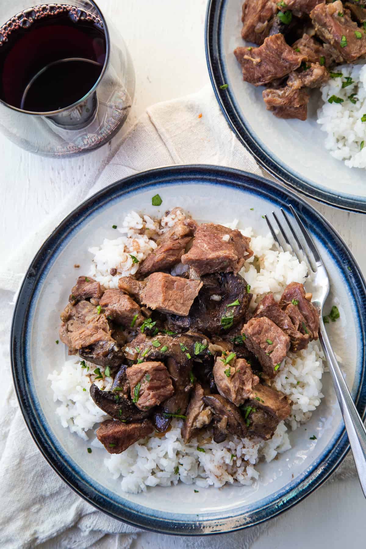 plate of beef stew on a bed of white rice on a blue rimmed plate, next to a glass of red wine.