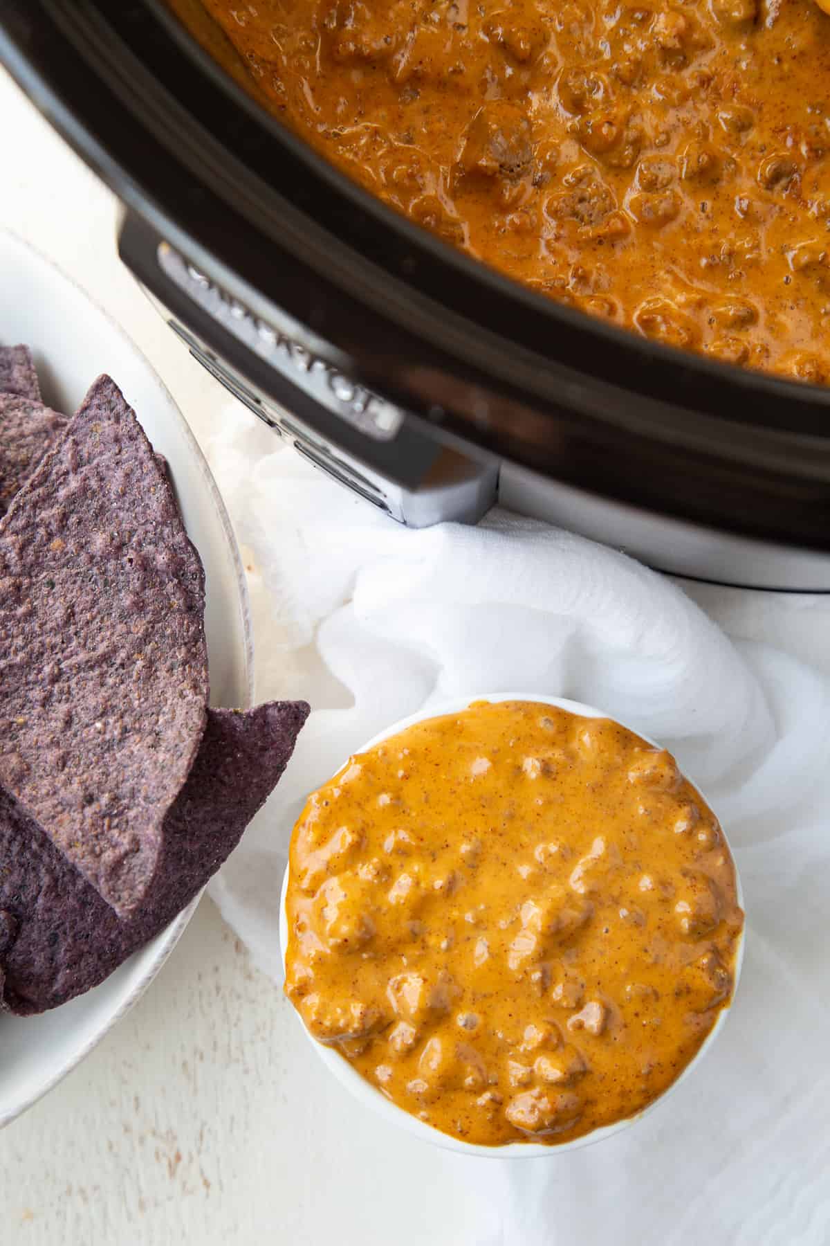 chili cheese dip in a ramekin next to a crockpot and a bowl of blue corn tortilla chips.