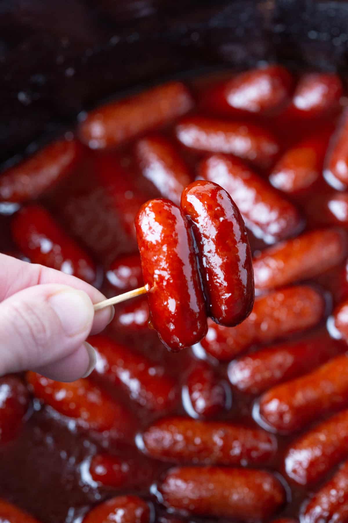 hand holding a toothpick with two little smokies hovering over a crockpot.