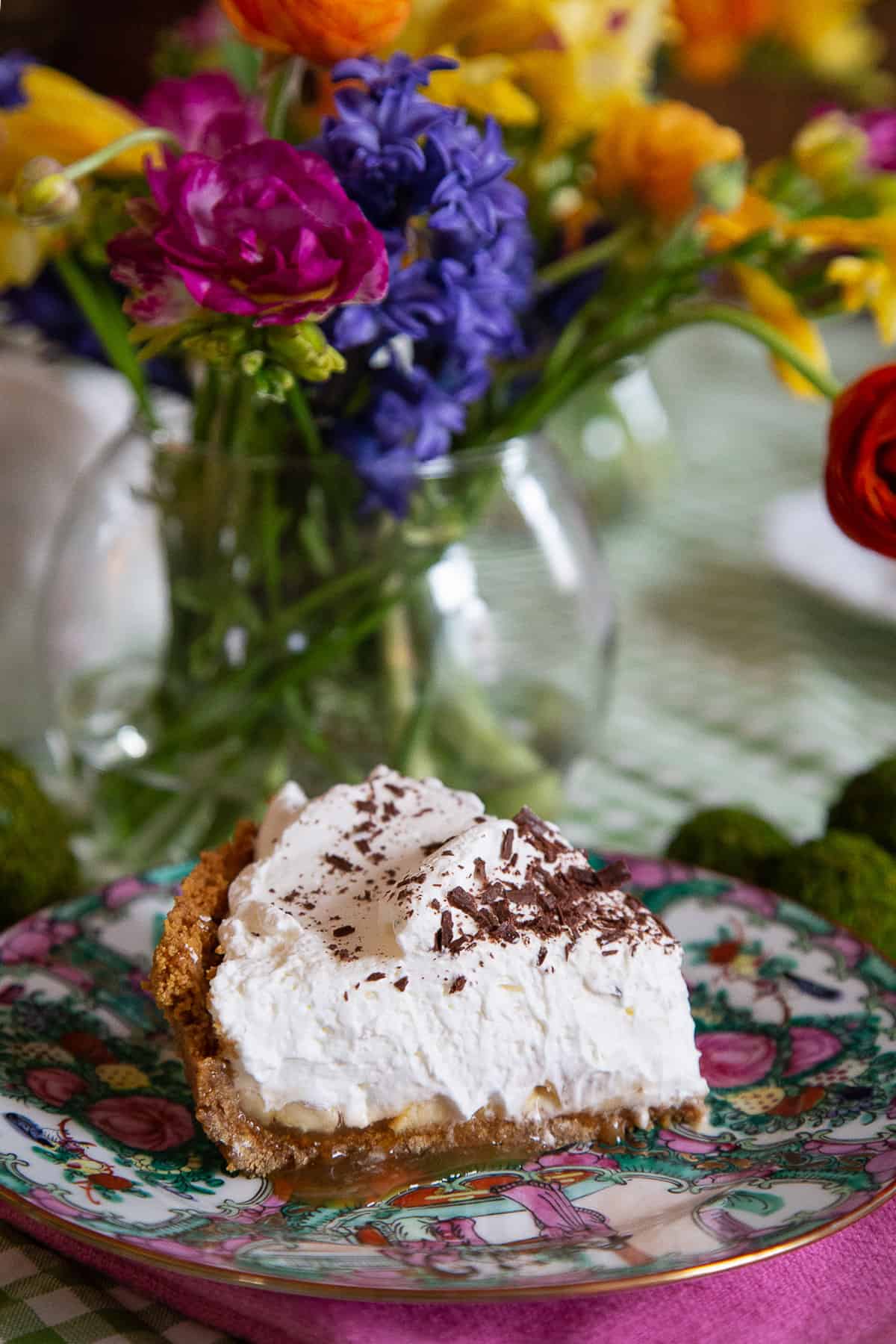 slice of banoffee pie on a china plate next to a vase of colorful flowers.