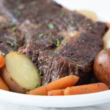 chuck roast made in the crockpot, sitting on a white platter with carrots and potatoes.