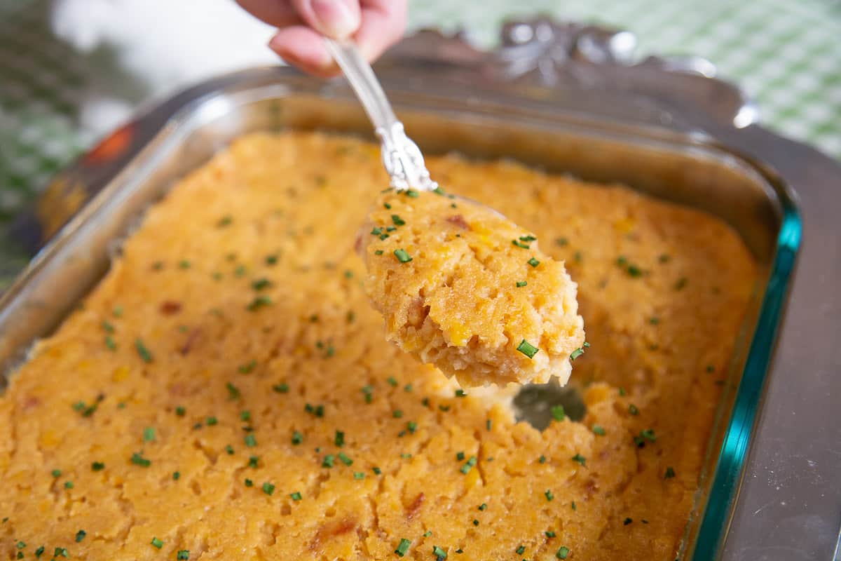 hand lifting scalloped corn with bacon out of a casserole dish.