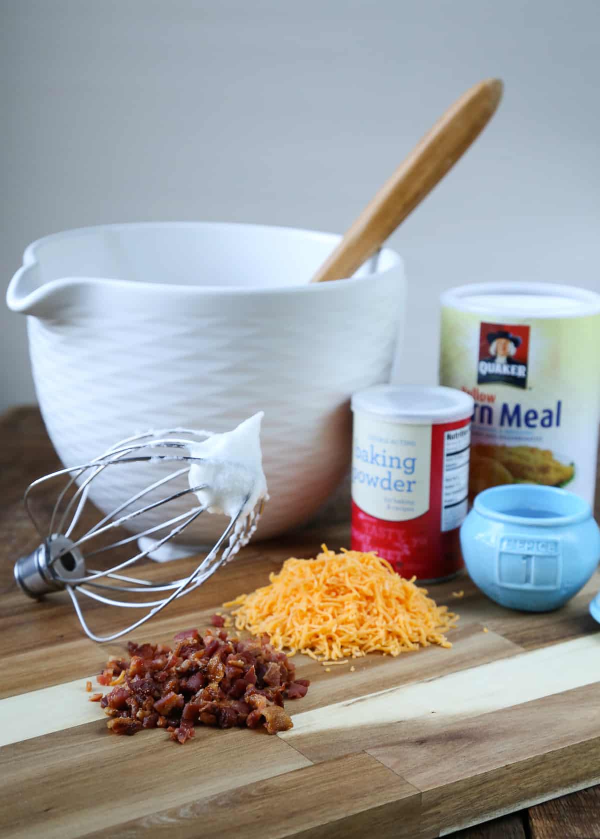 white mixing bowl, shredded cheddar, chopped bacon and other ingredients on a wooden board.