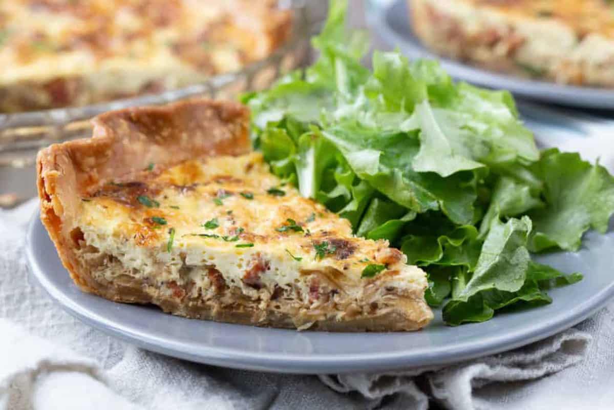 slice of alsatian quiche with bacon and caramelized onions on a gray plate with a leafy green salad.