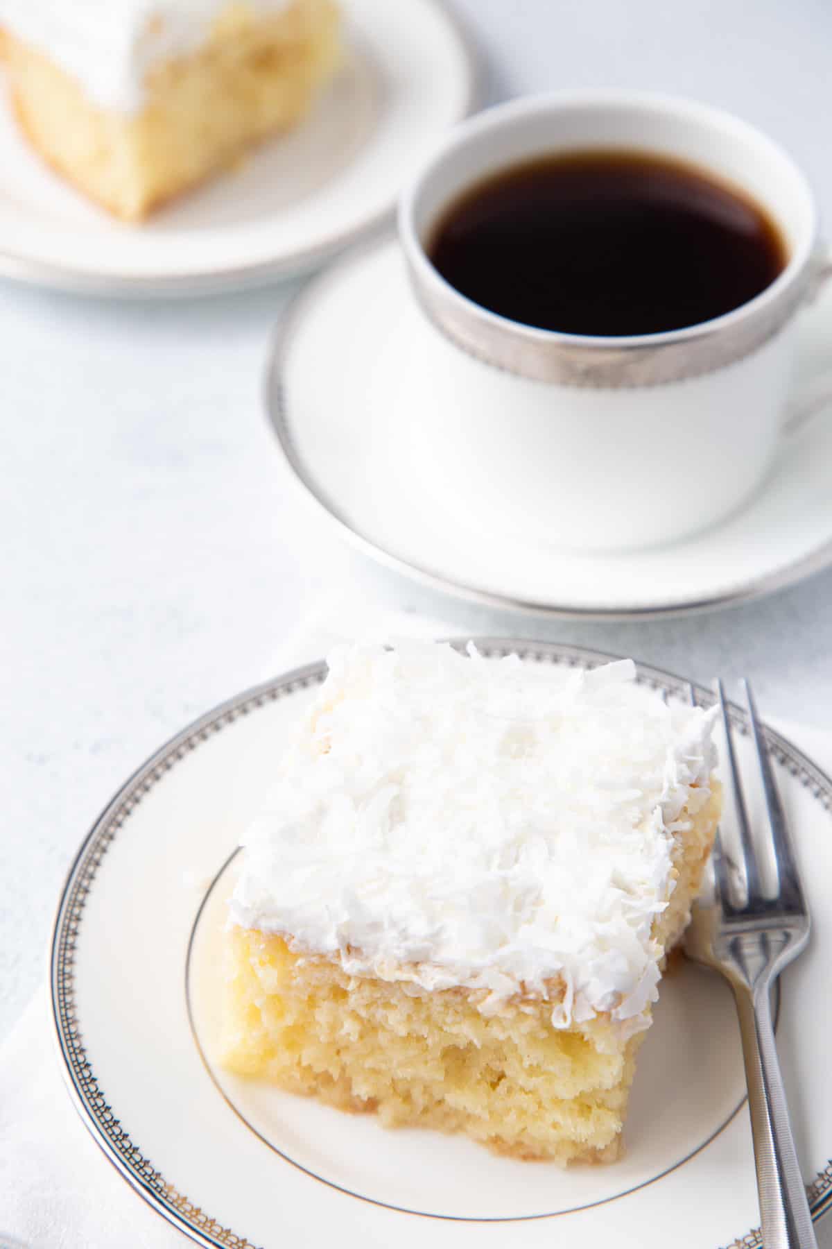 coconut poke cake on a white plate next to a mug of coffee.